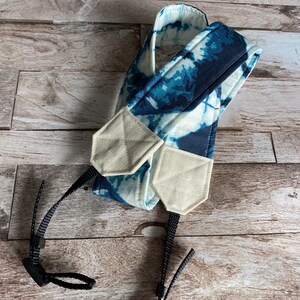 May include: A camera strap featuring a blue and white tie-dye design. The strap has beige fabric connectors and black adjustable nylon straps. The pattern is a mix of dark and light blue shades, offering a distinctive visual texture.