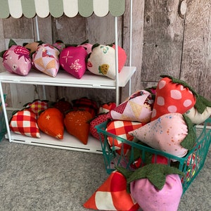 Puede incluir: Un peque&ntilde;o puesto de mercado blanco con un toldo a rayas verdes y blancas exhibe decoraciones de fresas de tela. Las fresas son de varios colores y patrones, incluyendo rosa, rojo y naranja, y est&aacute;n dispuestas en estantes y en una cesta verde.