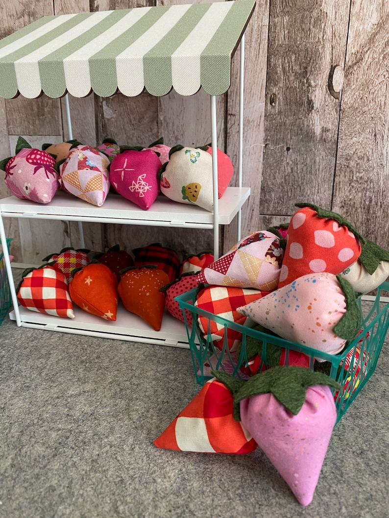 Puede incluir: Un peque&ntilde;o puesto de mercado blanco con un toldo a rayas verdes y blancas exhibe decoraciones de fresas de tela. Las fresas son de varios colores y patrones, incluyendo rosa, rojo y naranja, y est&aacute;n dispuestas en estantes y en una cesta verde.