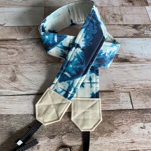 May include: A blue and white tie-dye camera strap with cream-coloured end pieces. The strap is made of fabric and has black adjustable straps for attaching to a camera. The design features shades of blue and white.