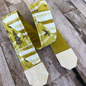 May include: A camera strap with a patterned design. The strap features a repeating pattern of birds, trees, and Japanese characters in shades of olive green, white, and brown. The strap ends are a solid olive green with off-white leather-like tabs.