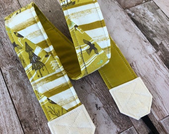 May include: A camera strap with a patterned design. The strap features a repeating pattern of birds, trees, and Japanese characters in shades of olive green, white, and brown. The strap ends are a solid olive green with off-white leather-like tabs.