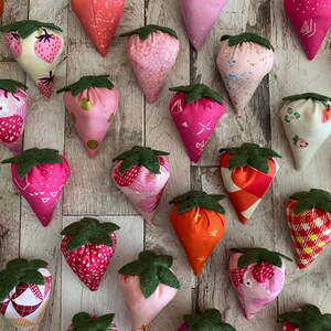 Puede incluir: Una colecci&oacute;n de adornos de fresas de tela en varios tonos de rosa y naranja, con hojas de fieltro verde. Las fresas presentan diferentes patrones, incluyendo lunares, estampados florales y dise&ntilde;os geom&eacute;tricos. El fondo es de vetas de madera clara.