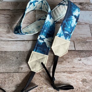 May include: A blue and white tie-dye camera strap with cream-coloured end pieces and black adjustable straps. The strap is made of fabric with a blue and white pattern. The end pieces are a neutral cream colour.