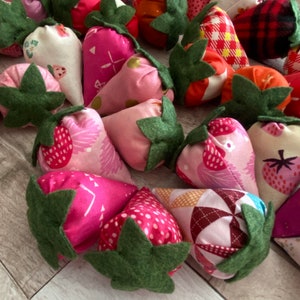 Puede incluir: Una colecci&oacute;n de fresas de tela hechas a mano en varios tonos de rosa, rojo y blanco. Cada fresa tiene una hoja de fieltro verde y est&aacute; hecha de tela estampada, incluyendo lunares, estampados florales y cuadros.