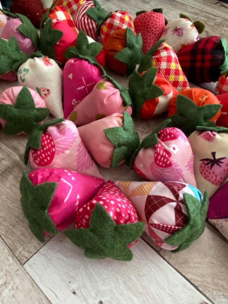 Puede incluir: Una colecci&oacute;n de fresas de tela hechas a mano en varios tonos de rosa, rojo y blanco. Cada fresa tiene una hoja de fieltro verde y est&aacute; hecha de tela estampada, incluyendo lunares, estampados florales y cuadros.