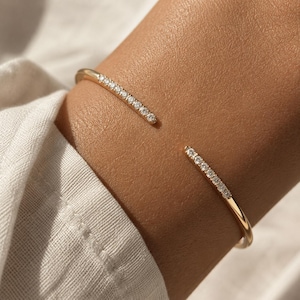 May include: A gold-colored open cuff bracelet with a row of small, clear gemstones set along each end. The bracelet is worn on a wrist, and the background is a soft, neutral color.