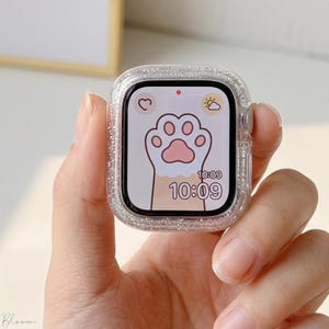Puede incluir: Una funda transparente para Apple Watch, cubierta de purpurina. La esfera del reloj muestra un diseño de huella de pata rosa con un corazón y un icono de sol. La hora es 10:09. La funda se sostiene en una mano.