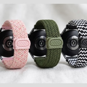 May include: Three smartwatch bands in a row. The first is pink with a matching clasp, the second is olive green, and the third is black and white zig-zag patterned. All three are attached to black smartwatches.