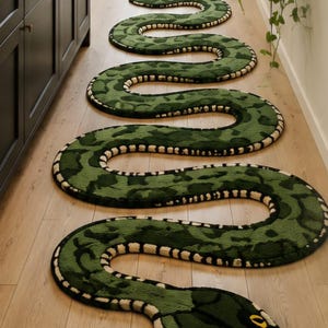 May include: A long, winding snake-shaped rug featuring a dark and light green camouflage-style pattern. The rug has a textured surface, a cream-colored underbelly border with black accents, and a head with two yellow eyes and a black forked tongue.