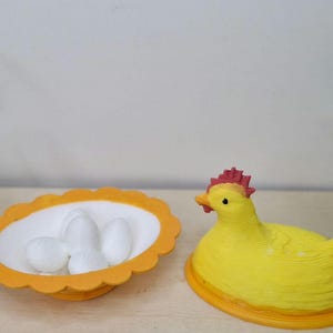 Può includere: Una statuetta di gallina in ceramica gialla con cresta rossa e base arancione. Accanto alla gallina c'è un piatto ovale arancione e bianco con tre uova bianche. Due piccole piante in vaso con foglie verdi su entrambi i lati.