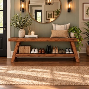 May include: A wooden entryway bench with a lower shelf holding a woven basket and various shoes. The top surface features a ceramic vase with greenery, a candle, books, and two decorative pillows. A round mirror hangs on the wall above.
