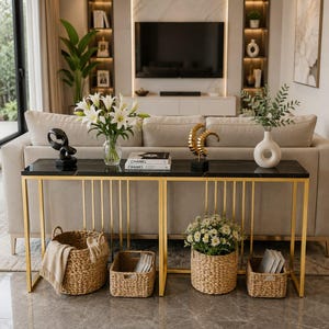 May include: A long console table with a black marble top and a gold metal frame. The table holds a glass vase with white lilies, two stacked Chanel books, a gold spiral sculpture, and a white ceramic vase with greenery. Four woven baskets sit below.