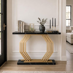 May include: A console table featuring a black marble top and base with a gold-toned metal frame in an octagonal geometric design. The table holds several books, a glass geometric terrarium, a dark gray vase with greenery, and three black candles.