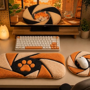 Puede incluir: Un reposamuñecas rectangular para teclado y una alfombrilla de ratón circular con un patrón texturizado y acolchado en naranja, crema y negro. El reposamuñecas muestra una huella de pata naranja y la alfombrilla sostiene un ratón blanco.