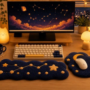 Könnte beinhalten: Eine marineblaue Tastatur-Handballenauflage und ein passendes Mauspad mit gelben getufteten Sternen und einem Sternbildmuster. Das Set liegt auf einem Holztisch mit einem Computermonitor, einer weißen kabellosen Maus und warmen Lampen.