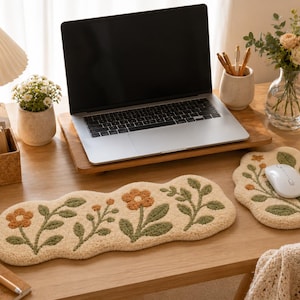 Puede incluir: Un reposamuñecas para teclado y una alfombrilla de ratón a juego, ambos con una base de color crema y un patrón floral y de hojas texturizado en tonos naranja y verde salvia. Los artículos tienen una forma orgánica irregular con bordes suaves.