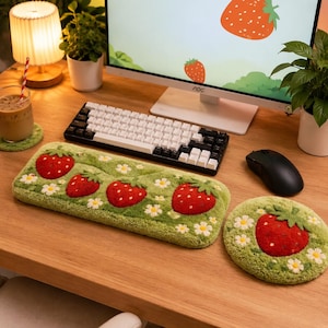 以下が含まれることがあります： 赤いイチゴと小さな白い花の模様が施された、緑色のテクスチャーのある表面を持つ長方形のキーボード用リストレストと円形のマウスパッド。木製のデスクの上に置かれ、コンピューターモニターの前に配置されています。