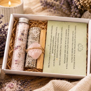 Può includere: Una scatola bianca piena di articoli per la cura di sé. Contiene una bottiglia di vetro di sali da bagno, mazzi di salvia, un cristallo di quarzo rosa e bastoncini di palo santo. È inclusa una carta con testo. La scatola è circondata da lavanda e una candela accesa.