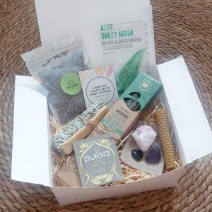 Könnte beinhalten: Eine weiße Box mit Pflegeprodukten, darunter eine Gesichtsmaske, Räucherstäbchen, Salbeibündel, Kristalle, eine Kerze und Tee. Die Box ist mit Stroh ausgekleidet und steht auf einer gewebten Matte. Die Artikel sind in verschiedenen Farben gehalten.