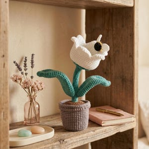 Può includere: Un fiore bianco all'uncinetto con centro nero e foglie verdi in un vaso grigio. Il fiore fatto a mano è su una mensola di legno con un piccolo vaso di fiori secchi, un taccuino e una penna. La mensola è in legno stagionato.