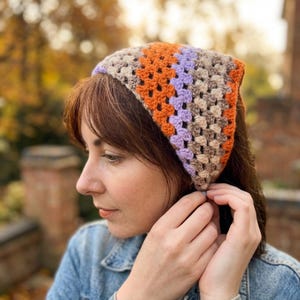 Puede incluir: Un pañuelo para la cabeza de ganchillo colorido con un patrón de cuadrados de la abuela. El pañuelo presenta cuadrados naranjas, lavanda y beige. La prenda para la cabeza tiene forma triangular y se lleva en la cabeza.