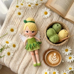 May include: A handmade crochet fairy doll with blonde hair, green dress, and white wings. The doll is on a cream-colored knitted blanket, surrounded by daisies. A basket of green and yellow yarn, a crochet hook, and a cup of coffee are also present.