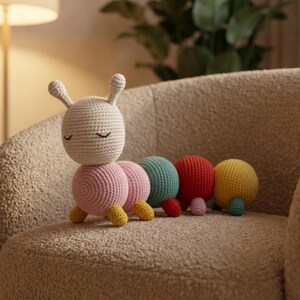 May include: A handmade crochet caterpillar toy with a cream-colored head, pink body segments, and colorful segments of teal, red, and yellow. The caterpillar has closed eyes and small yellow feet. The toy is sitting on a beige armchair.