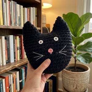 May include: A black crocheted cat-shaped pillow with white eyes, white whiskers, and a pink nose. The handmade cat pillow is held in front of a bookshelf and a potted plant.