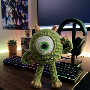 May include: A handmade green crochet monster with one large eye, small horns, and arms raised in a flex. The plush toy is on a wooden desk near a keyboard, headphones, and a computer monitor. The monster is a character from the movie Monsters, Inc.