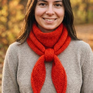 May include: A woman wearing a bright red knitted scarf tied in a knot. The scarf is made of a thick yarn and has a textured appearance. The woman is wearing a gray sweater, and the background is blurred with autumn colors.