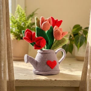 Könnte beinhalten: Ein gehäkeltes Tulpenbouquet in einer lavendelfarbenen Gießkanne. Die Tulpen sind rot und pfirsichfarben, mit grünen Blättern. Ein rotes Herz ist an der Gießkanne befestigt. Das Arrangement steht auf einer weißen Fensterbank, mit Pflanzen im Hintergrund.
