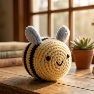 May include: A handmade crochet bee with a yellow body, black stripes, and light blue wings. The bee has a smiling face with black eyes and a curved mouth. The plush toy is sitting on a wooden surface.