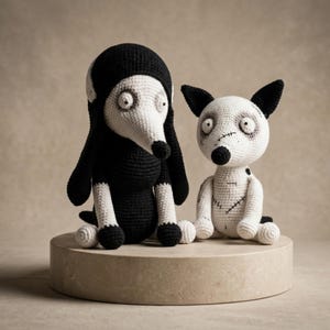 May include: Two handmade crochet dog dolls. One is black and white, wearing a black hood. The other is white with black ears and stitched details. Both have black noses and large, round eyes. They sit on a light-colored, square pedestal.