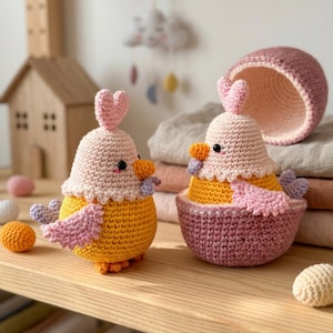 May include: Two handmade crochet chick figurines. One chick is standing, the other is inside a pink basket. Both have yellow bodies, pink wings, and heart-shaped headpieces. The chicks are on a wooden surface.