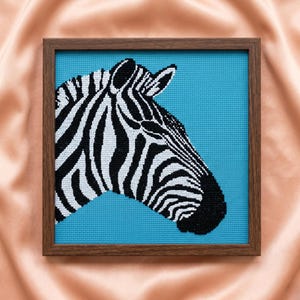 Peut inclure: Une broderie au point de croix carrée représentant le profil d'un zèbre aux rayures noires et blanches sur un fond turquoise vif, encadrée dans un cadre en bois brun foncé simple avec une texture de grille visible sur le tissu.