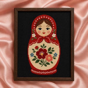 Può includere: Un ricamo a punto croce incorniciato raffigurante una bambola russa su sfondo nero. La bambola indossa un foulard rosso a pois bianchi e un grembiule color crema decorato con un motivo centrale di un bouquet di fiori rossi e rosa.