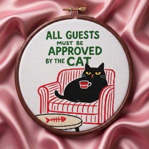 Puede incluir: Un bastidor de punto de cruz con un gato negro en un sillón a rayas rojas, sosteniendo una taza. El texto dice "ALL GUESTS MUST BE APPROVED BY THE CAT". Una pequeña mesa con un diseño de espina de pescado está frente al gato.