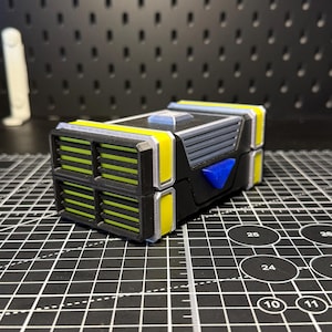 May include: A rectangular, multi-colored plastic container featuring a black base, bright yellow accents, and silver trim. The front panel displays a grid of horizontal slats in lime green, while a small, triangular blue piece is attached to the side.