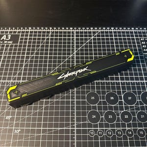 May include: A long, rectangular black plastic case featuring the Cyberpunk logo in white text. The case has bright yellow accents on both ends, each marked with a small plus sign. The surface has a textured, grooved pattern along the center.