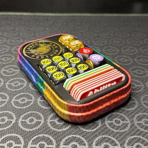 May include: A rectangular multi-coloured storage case containing a black circular 2025 Pokemon Day coin, several black dice marked with the number 100, yellow dice with GX symbols, and a set of white and red plastic markers labelled Ability.