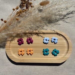 Peut inclure: Six boucles d'oreilles en forme de fleurs de couleurs violet, bleu clair, orange et turquoise sont présentées sur un plateau en bois. Les boucles d'oreilles sont disposées par paires. Des graminées ornementales séchées et des capsules de graines sont en arrière-plan.