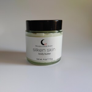 May include: A clear glass jar with a black lid, containing body butter. The jar has a white label with the text "silken skin body butter" and the brand name "WICKED GOOD BODY". The net weight is 4 oz / 113g.