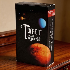 May include: A black tarot card deck with the words "Tarot with Keywords" in white gothic font. The box features a planetary design with a blue planet and a reddish-orange planet. The side of the box reads "78 Tarot Cards Deck for Beginners".