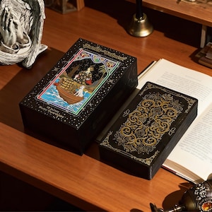 May include: A black tarot card box with gold lettering and a colourful illustration of a boat with a person and a dog. The box is open, revealing a deck of cards with gold filigree on a black background. The text "Tarot-Waite" is visible.