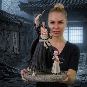 May include: A detailed anime-style figurine set featuring a tall, muscular male character with spiky black hair wearing a black robe and a smaller female character with pink hair in a light-colored dress, both standing on a textured circular base.