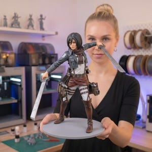 May include: A detailed figurine of an Attack on Titan character, holding two swords. The figure wears a gray jacket, brown boots, and a white shirt. The figure stands on a grey circular base. The figure is approximately 20 cm tall.