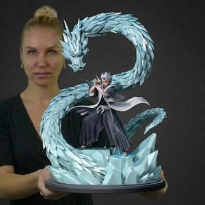Peut inclure: Une figurine de style anime détaillée représentant un personnage aux cheveux en pointe vêtu d'un kimono sombre, dans une pose dynamique. La figurine est entourée d'un grand dragon de glace bleu clair translucide aux écailles cristallines.