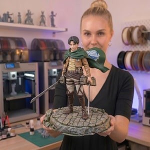 May include: A detailed figurine of a character from the anime series Attack on Titan. The figure is dressed in a brown jacket, tan trousers, and tall brown boots, with a green cape. It is holding two swords and is mounted on a stone-like base.