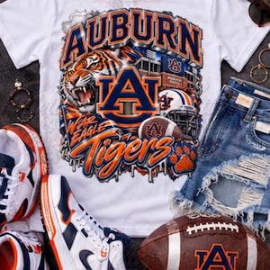 May include: A white short-sleeved t-shirt featuring a graphic of a tiger, a football helmet, a football, and the text Auburn Tigers and Wear Eagle. The shirt is displayed with blue denim shorts, white, orange, and navy trainers, and a football.